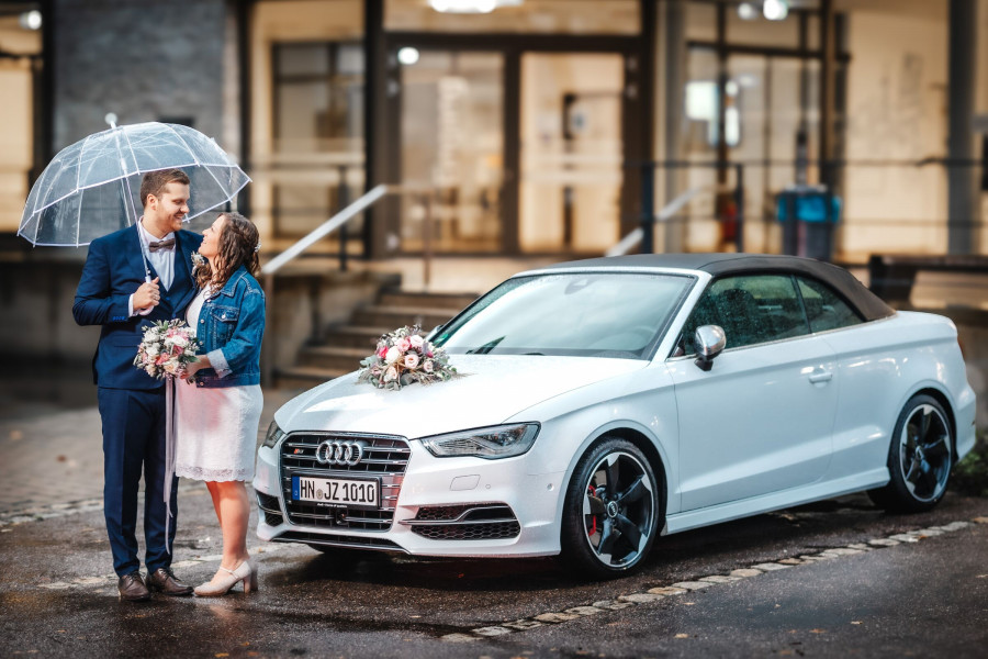Brautpaar im Regen neben geschmuecktem Hochzeitsauto